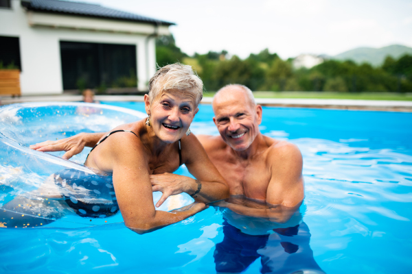 Portrait of cheerful senior couple in swimming pool outdoors in backyard, looking at camera.