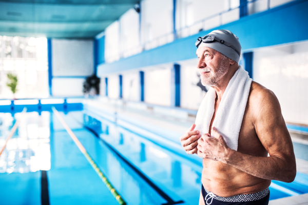 Senior man standing by the indoor swimming pool, a towel around his neck. Active pensioner enjoying sport.