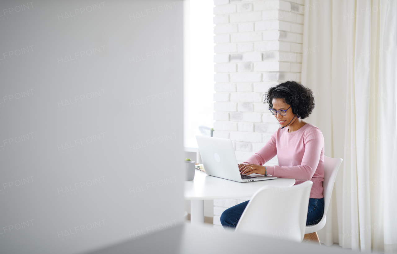 A portrait of happy mature woman with headset working on laptop indoors, home office concept.