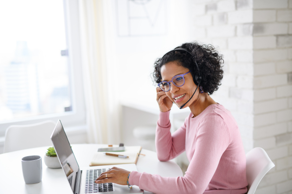 A portrait of happy mature woman with headset working on laptop indoors, home office concept.