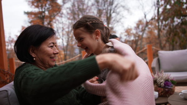 A happy little girl with grandmother sitting together wrapped in blanket outdoors in patio in autumn