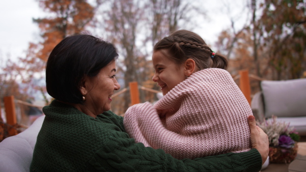 A happy little girl with grandmother sitting wrapped in blanket outdoors in patio in autumn