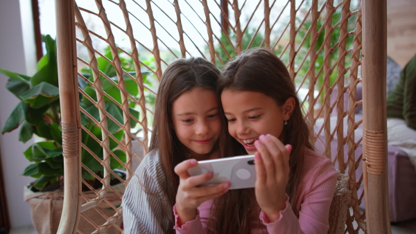 Happy little sisters sitting in a wicker rattan hang chair and using smartphone indoors in conservatory.