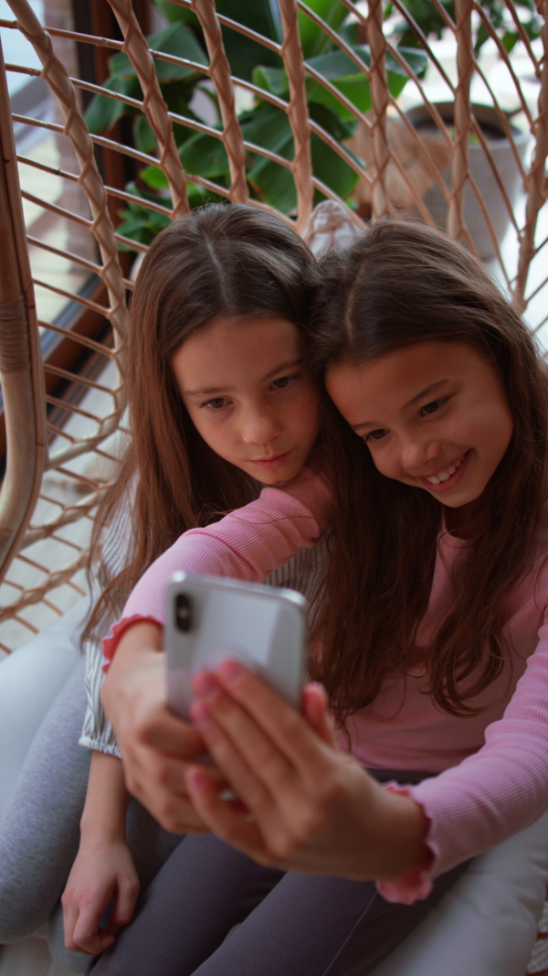 Happy little sisters sitting in a wicker rattan hang chair indoors in conservatory and taking selfie.