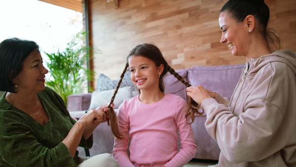 A happy little girl sitting on floor, her mother and grandmother making her plaits at home.