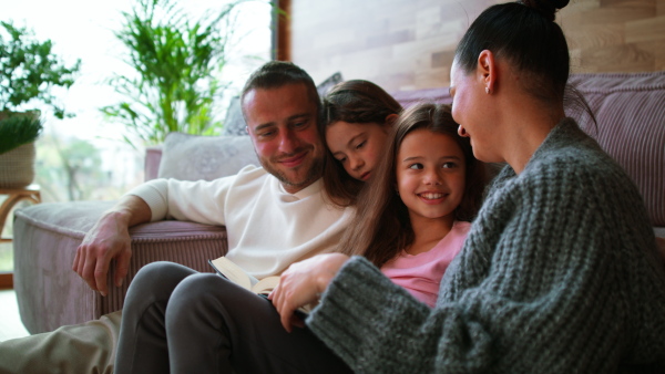 A young family with two little children sitting on floor and reading book at home.