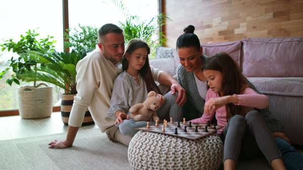 Two happy sisters with a mother and father sitting on floor and playing chess together at home