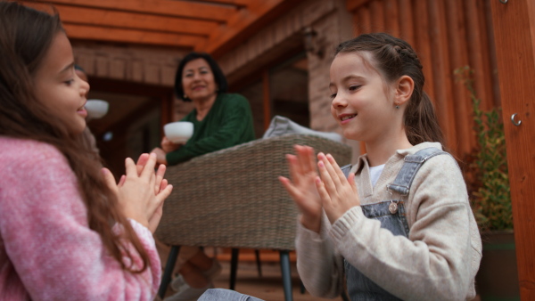 Two happy little sisters playing a clap hand game outdoors in patio in autumn, mother and grandmother at background.