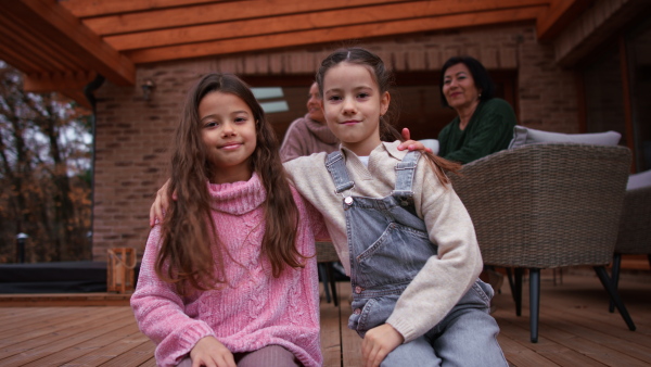 Two happy little sisters sitting with arms around outdoors in patio in autumn, mother and grandmother at background.