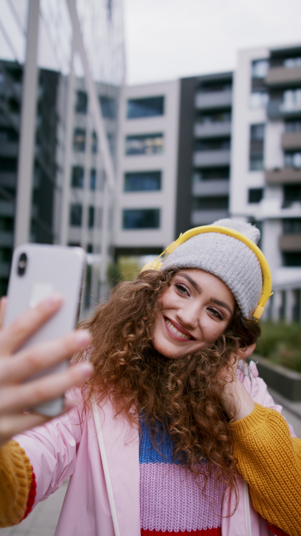 Young woman with hat standing outdoors on street, taking selfie, vertical video.