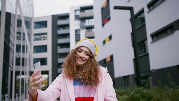 Young woman with hat standing outdoors on street, taking selfie.