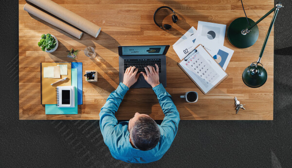 A top view of businessman working on laptop computer at desk with paperwork in home office.