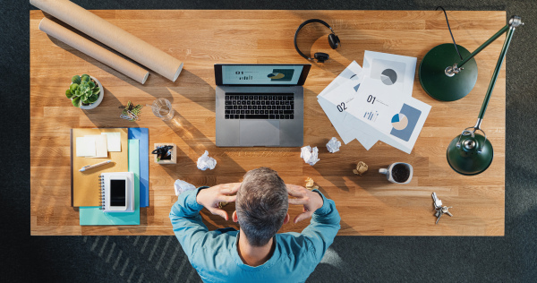 Top view of tired and frustrated businessman working on computer at desk with paperwork in home office.