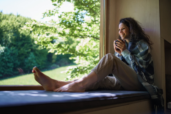 A happy woman with coffee sitting indoors in a tree house, weekend away and digital detox concept.