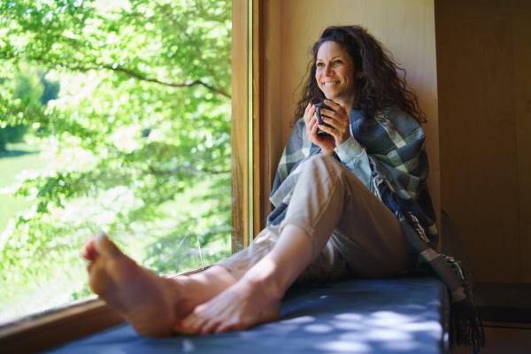 A happy mid adult woman with coffee sitting indoors in a tree house, weekend away and digital detox concept.