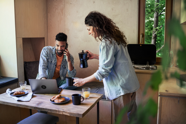 A happy couple with laptop sitting, resting and talking indoors in a tree house, remote office concept.