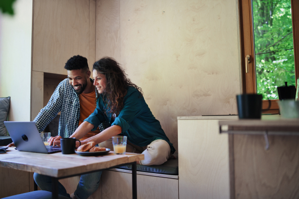 A happy couple with laptop sitting, resting and talking indoors in a tree house, remote office concept.