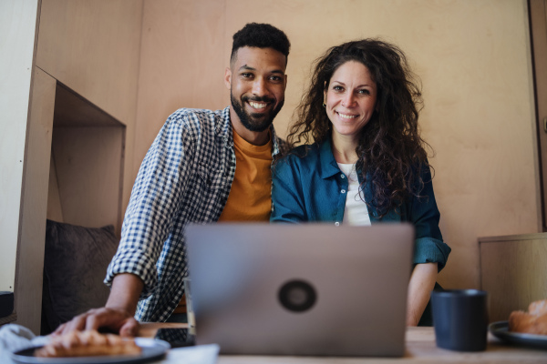 A happy biracial couple with laptop sitting, resting and looking at camera indoors in a tree house, weekend away and remote office concept.