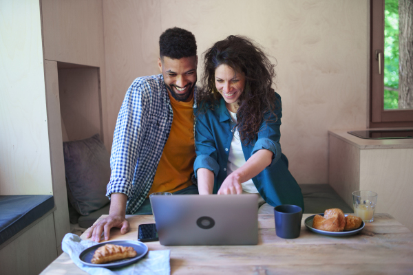 A happy couple with laptop sitting, resting and talking indoors in a tree house, weekend away and remote office concept.