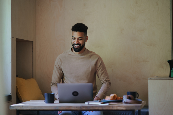 A happy young man with laptop and smartphone sitting, resting indoors in a tree house, weekend away and remote office concept.