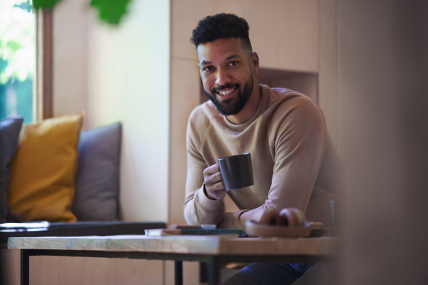 A happy young man with coffee sitting indoors in a tree house, weekend away and individual traveling concept.