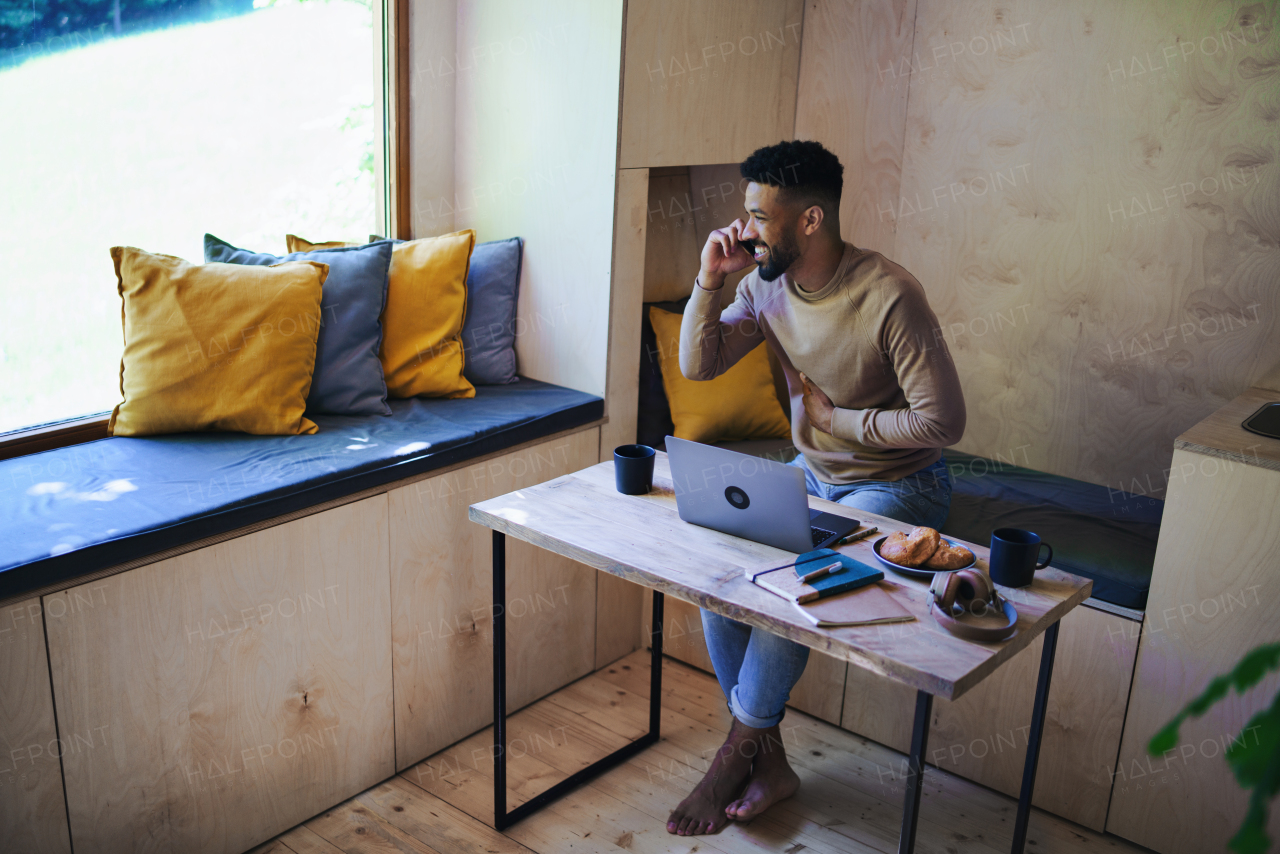 A happy young man with laptop and smartphone sitting, resting indoors in a tree house, weekend away and and remote office concept.