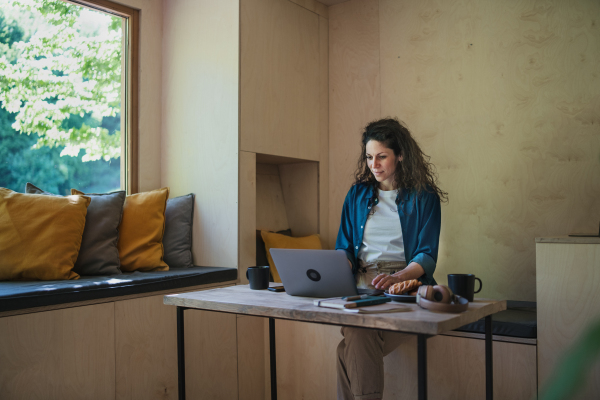 A happy young woman with coffee sitting and working on laptop indoors in a tree house, weekend away and individual traveling concept.