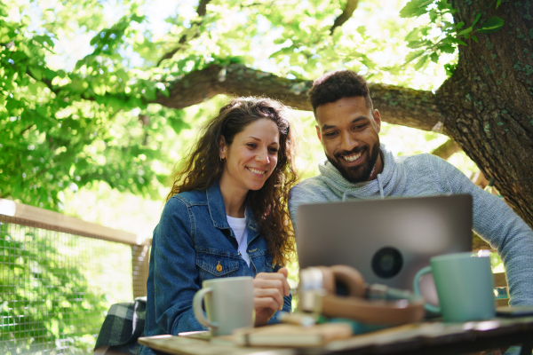 A happy couple with laptop resting outdoors in a tree house, weekend away and remote office concept.