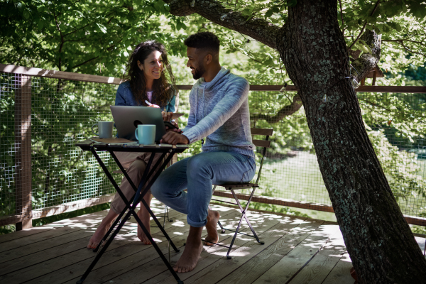 A happy couple with laptop resting outdoors in a tree house, weekend away and remote office concept.