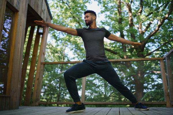 A low angle view of happy young man doing exercise outdoors on terrace of tree house, weekend away and digital detox concept.