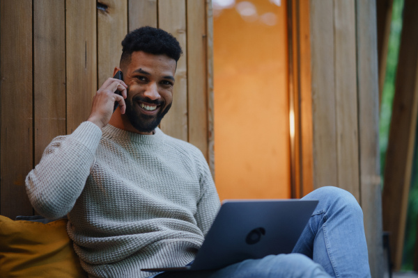 A happy young man with laptop and smartphone resting outdoors in a tree house, weekend away and remote office concept