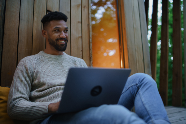 A happy young man with laptop resting outdoors in a tree house, weekend away and remote office concept.