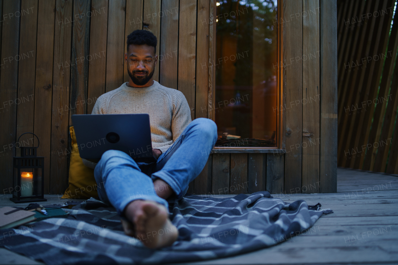 A happy young man with laptop resting outdoors in a tree house, weekend away and remote office concept.