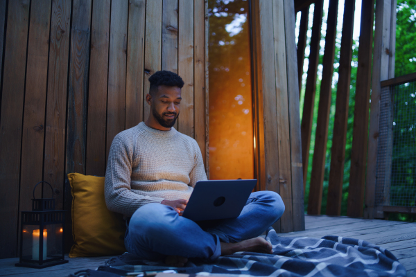 A happy young man with laptop resting outdoors in a tree house, weekend away and remote office concept.