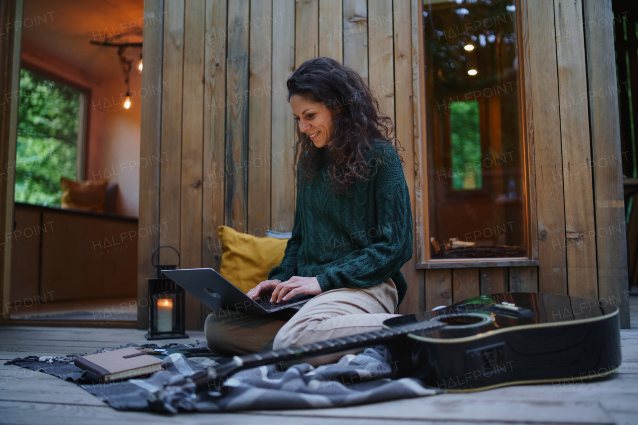 A happy young woman with laptop resting outdoors in a tree house, weekend away and remote office concept.