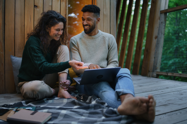 A happy couple with laptop resting outdoors in a tree house, weekend away and remote office concept.