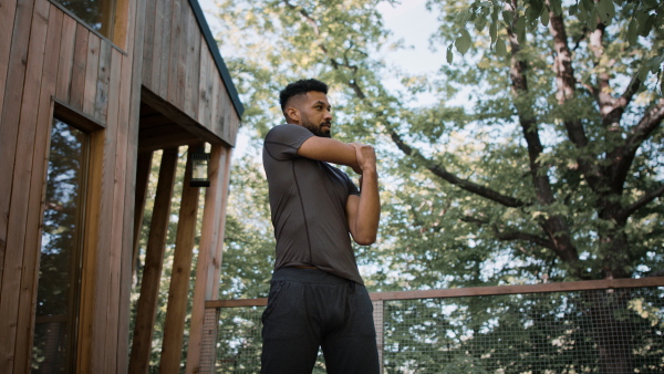 A low angle view of happy young man doing exercise outdoors on terrace of tree house, weekend away and digital detox concept.