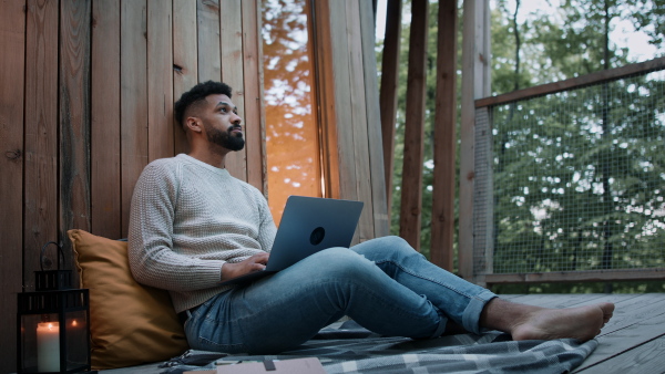 A happy young man with laptop getting call outdoors in a tree house, weekend away and remote office concept.