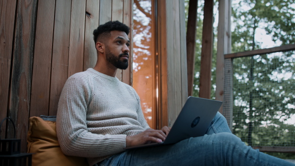 A happy young man with laptop getting call outdoors in a tree house, weekend away and remote office concept.