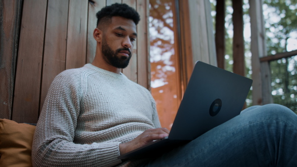 A happy young man with laptop resting outdoors in a tree house, weekend away and remote office concept.