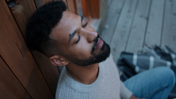 A close up of young man with closed eyes sitting outdoors on terrace of tree house, relaxing. Weekend away and digital detox concept.