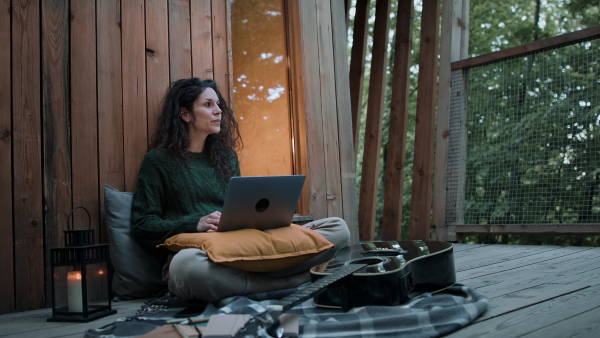 A happy young woman with laptop resting outdoors in a tree house, weekend away and remote office concept.