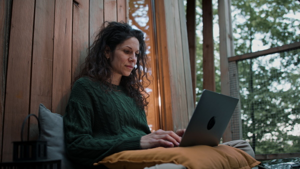 A happy young woman with laptop resting outdoors in a tree house, weekend away and remote office concept.