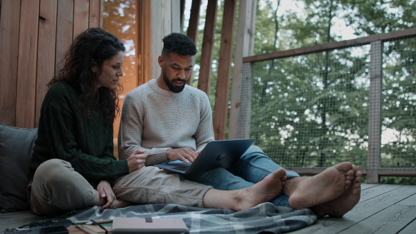 A happy couple with laptop resting outdoors in a tree house, weekend away and remote office concept.