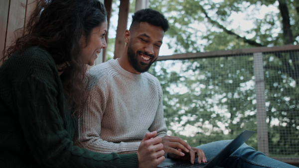 A happy couple with laptop resting outdoors in a tree house, weekend away and remote office concept.