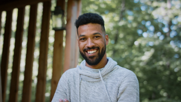 A happy young man standing outdoors on terrace of tree house, looking at camera. Weekend away and digital detox concept.