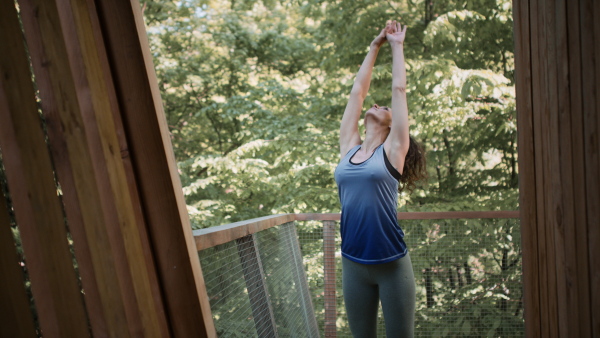 A happy mid adult woman doing exercise outdoors on terrace of tree house, weekend away and digital detox concept.