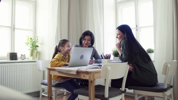 A happy small girl with mother and mother sitting at table at home, using laptop.