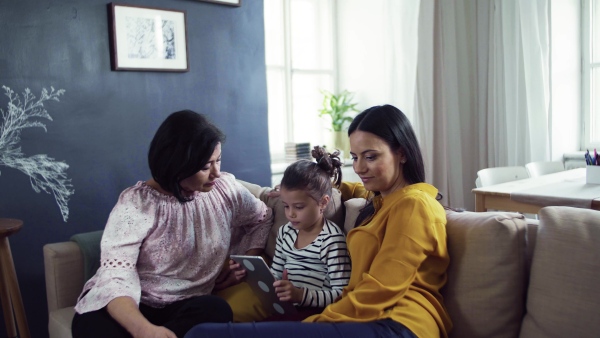 A happy small girl with mother and grandmother sitting on sofa at home, using tablet.