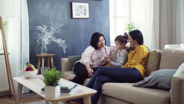 A happy small girl with mother and grandmother sitting on sofa at home, using tablet.